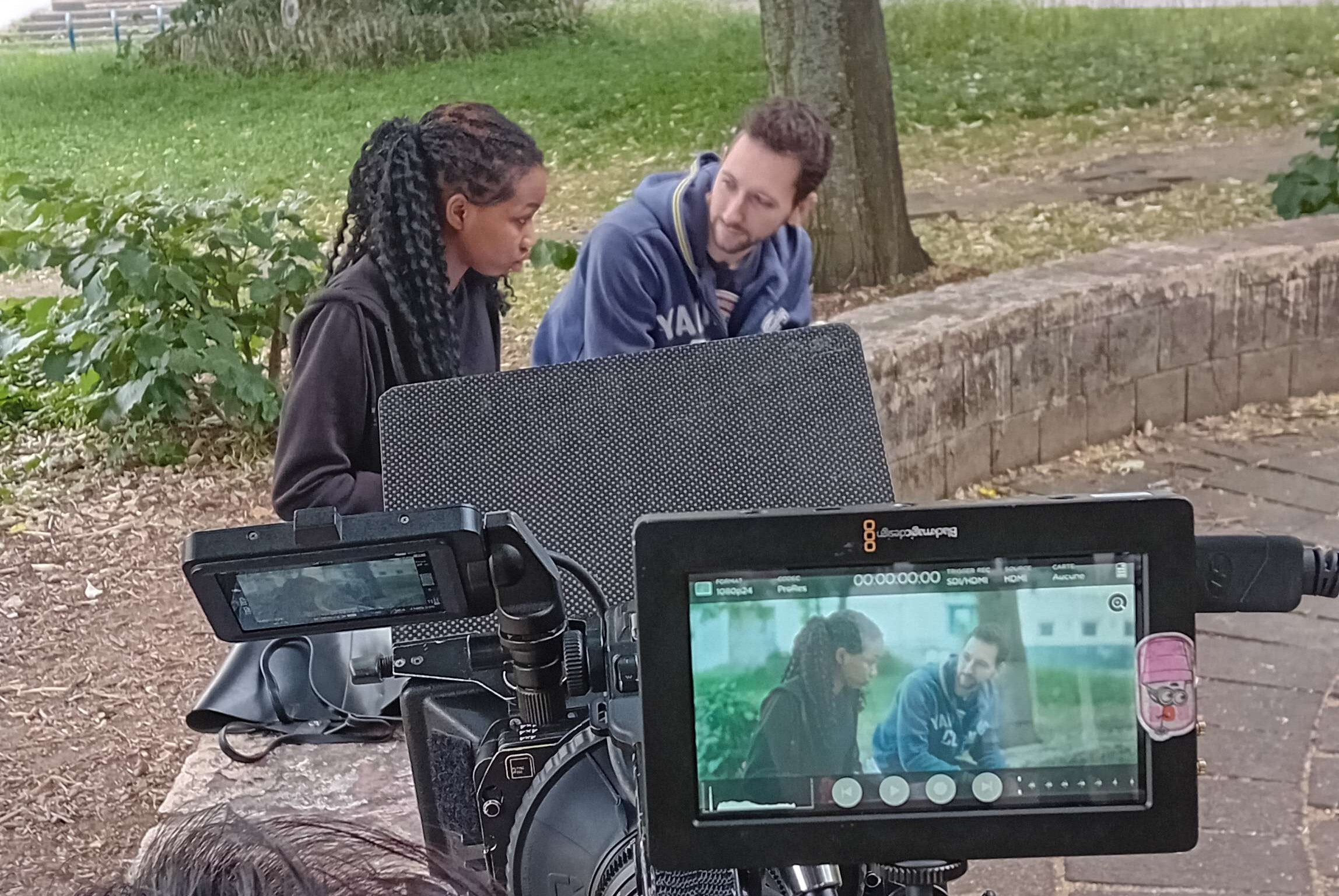 Vue sur une caméra portée à l'épaule filmant un couple assis sur un banc de pierre en extérieur
